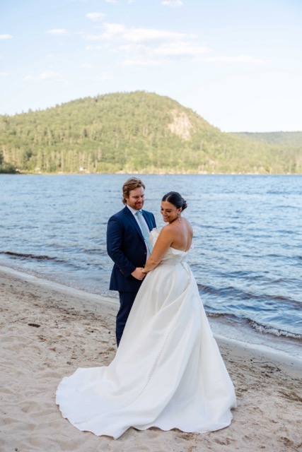 Bride and groom embracing on the sandy shore of Bear Lake at Camp Wigwam in Waterford, Maine, with the forested mountains of western Maine in the background