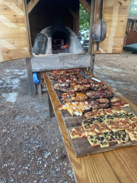 Wood-fired pizza oven at Camp Wigwam with a long board of fresh-baked artisan pizzas, used for rehearsal dinners and casual catering