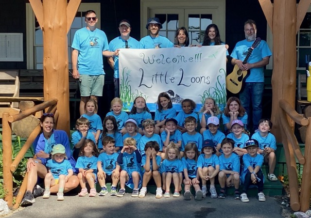 Group photo of Little Loons campers and staff holding a hand-painted Welcome Little Loons banner on the Wigwam Lodge porch