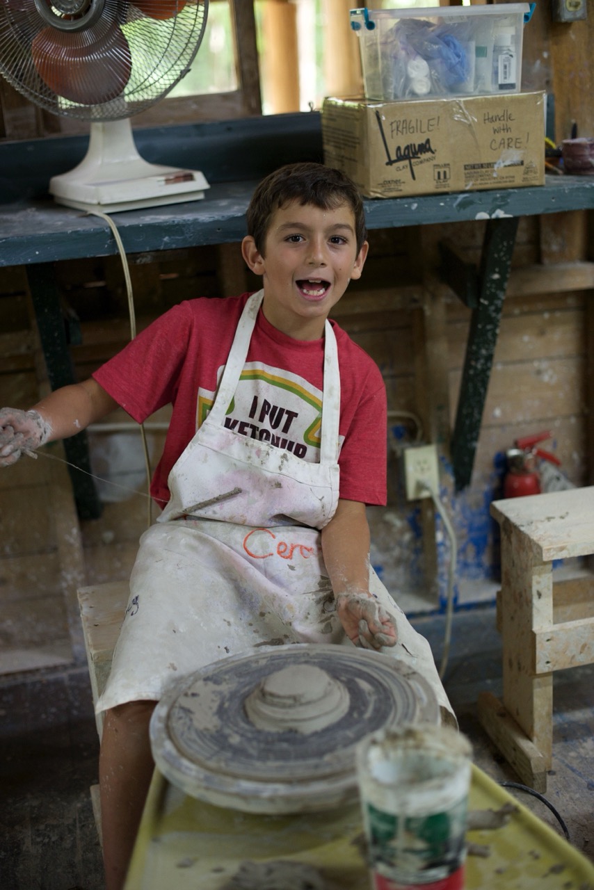 Smiling camper in a clay-covered apron sitting at the pottery wheel in the ceramics studio