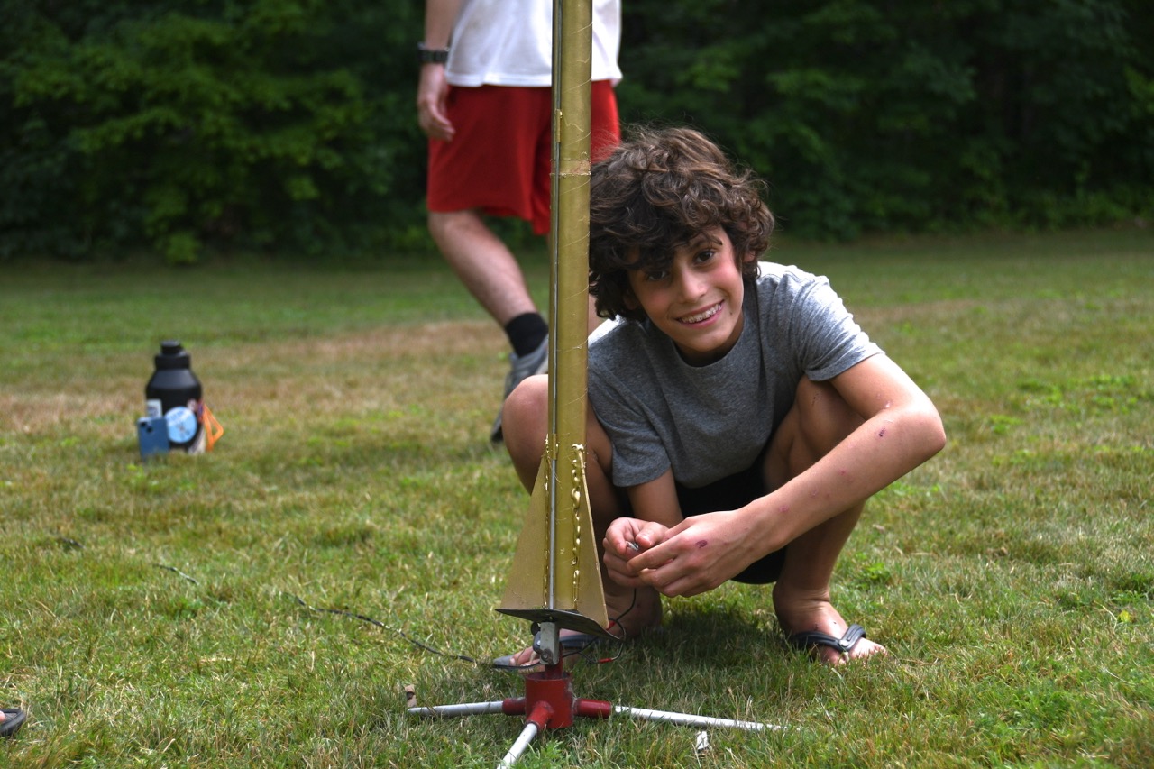 Camper kneeling on the grass beside a tall handmade rocket ready for launch at rocketry
