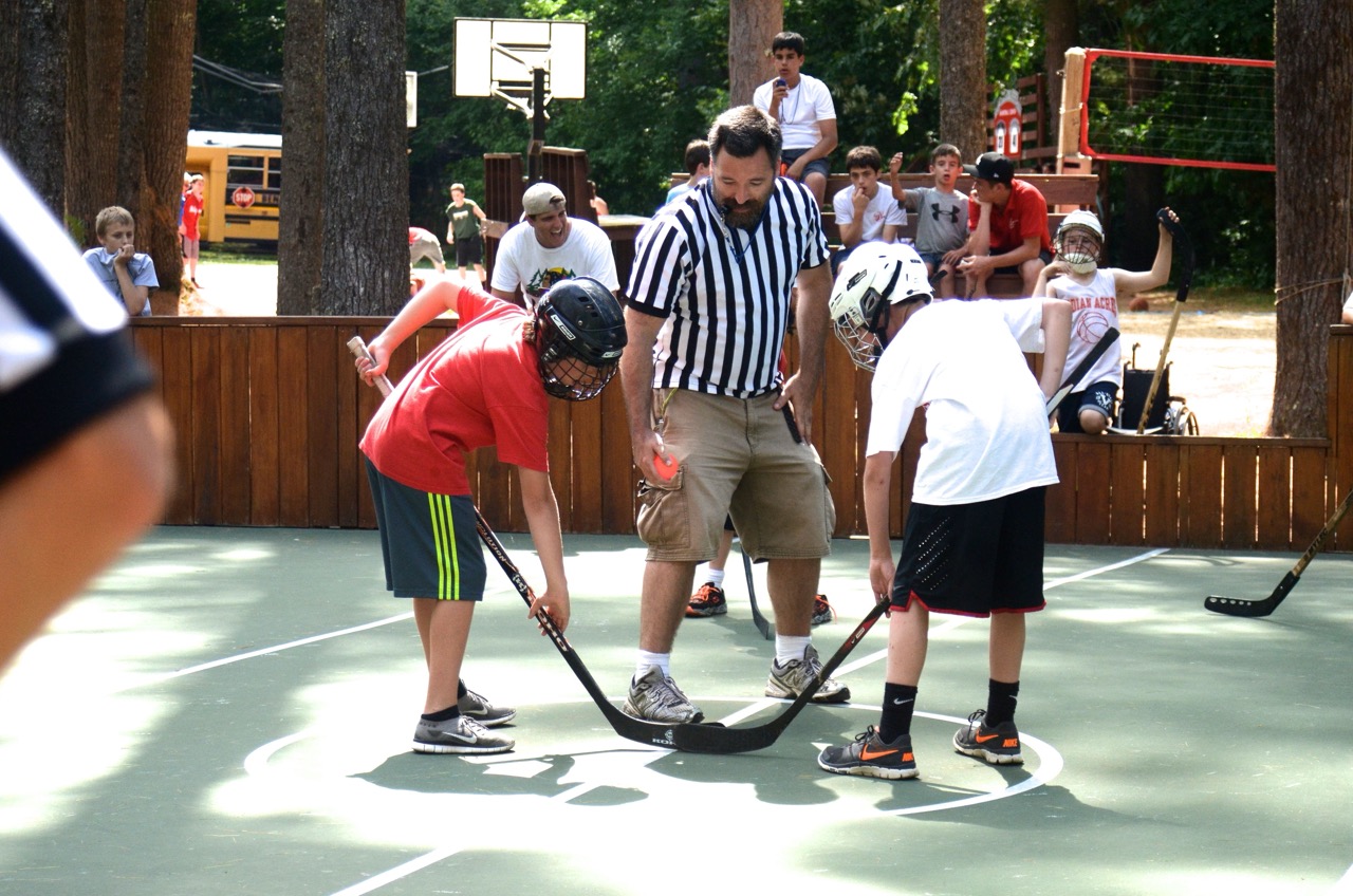 Referee dropping the ball between two helmeted campers for a face-off during a street hockey game
