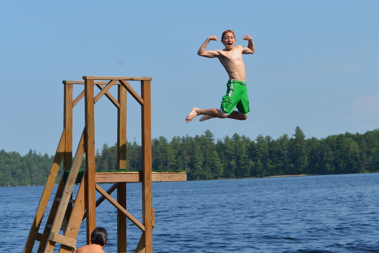 Camper flexing mid-air after leaping from the waterfront jump tower into Bear Lake