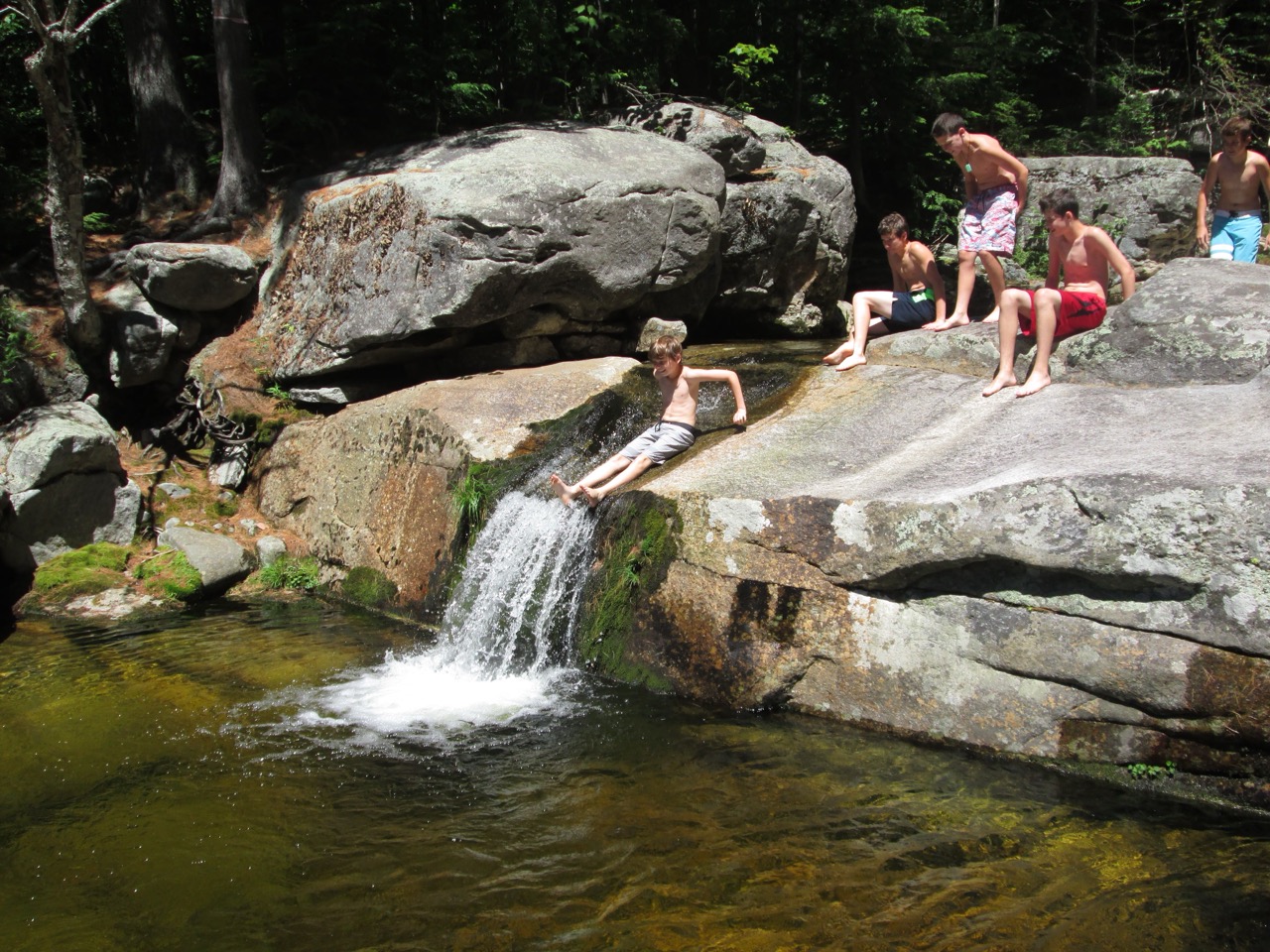 Camper sliding down a natural granite waterfall into a clear swimming hole on a camp hiking trip