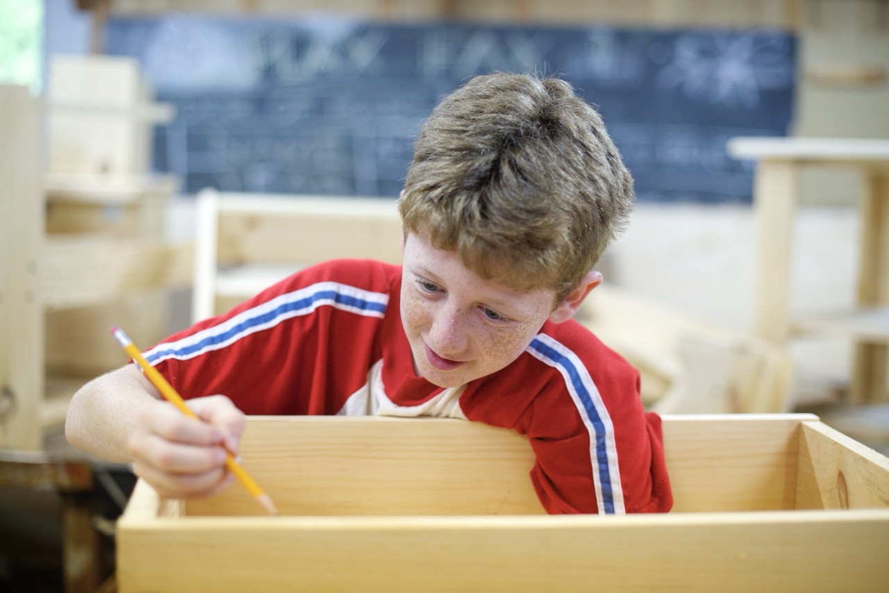 Focused camper sketching a design on a handmade wooden box in the woodworking shop
